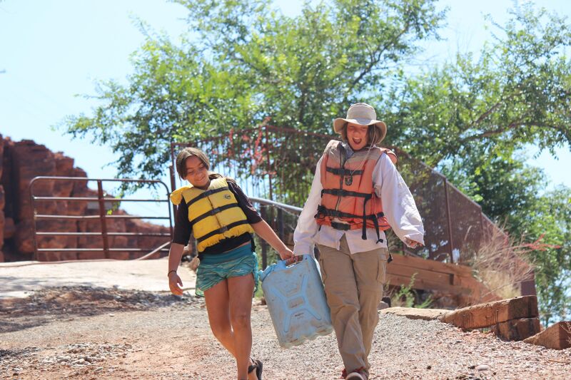 Two people, likely a parent and child, are walking on a path near a river or lake. Both are wearing life jackets, suggesting they may have been rafting or boating. The child is carrying a blue bag, and the adult is wearing a hat. The background shows trees and what appears to be a rocky landscape. They seem to be enjoying a day outdoors, possibly after a water activity.
